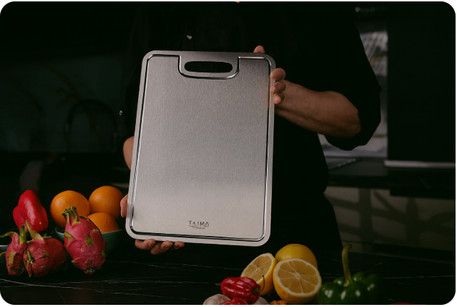 Person holding a titanium cutting board with handle in a modern kitchen, surrounded by fresh fruits and vegetables.